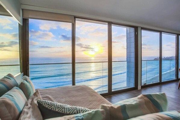 A room with a large window overlooking the ocean, a sofa with cushions, and a view of the sunset.