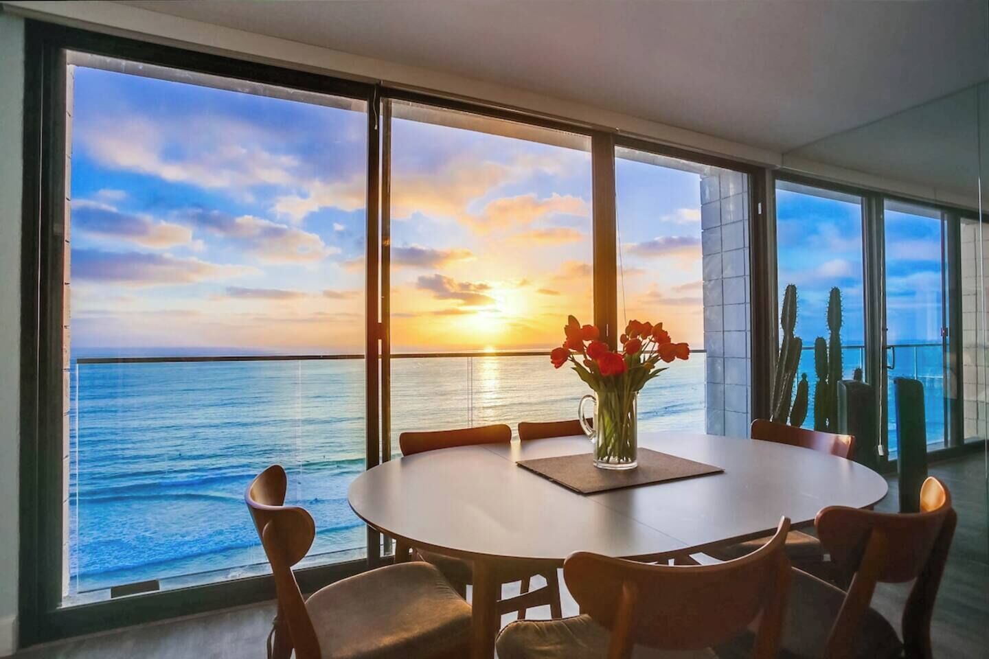 Un comedor con mesa y sillas, un jarrón con flores y vista al océano y al atardecer a través de amplias ventanas.