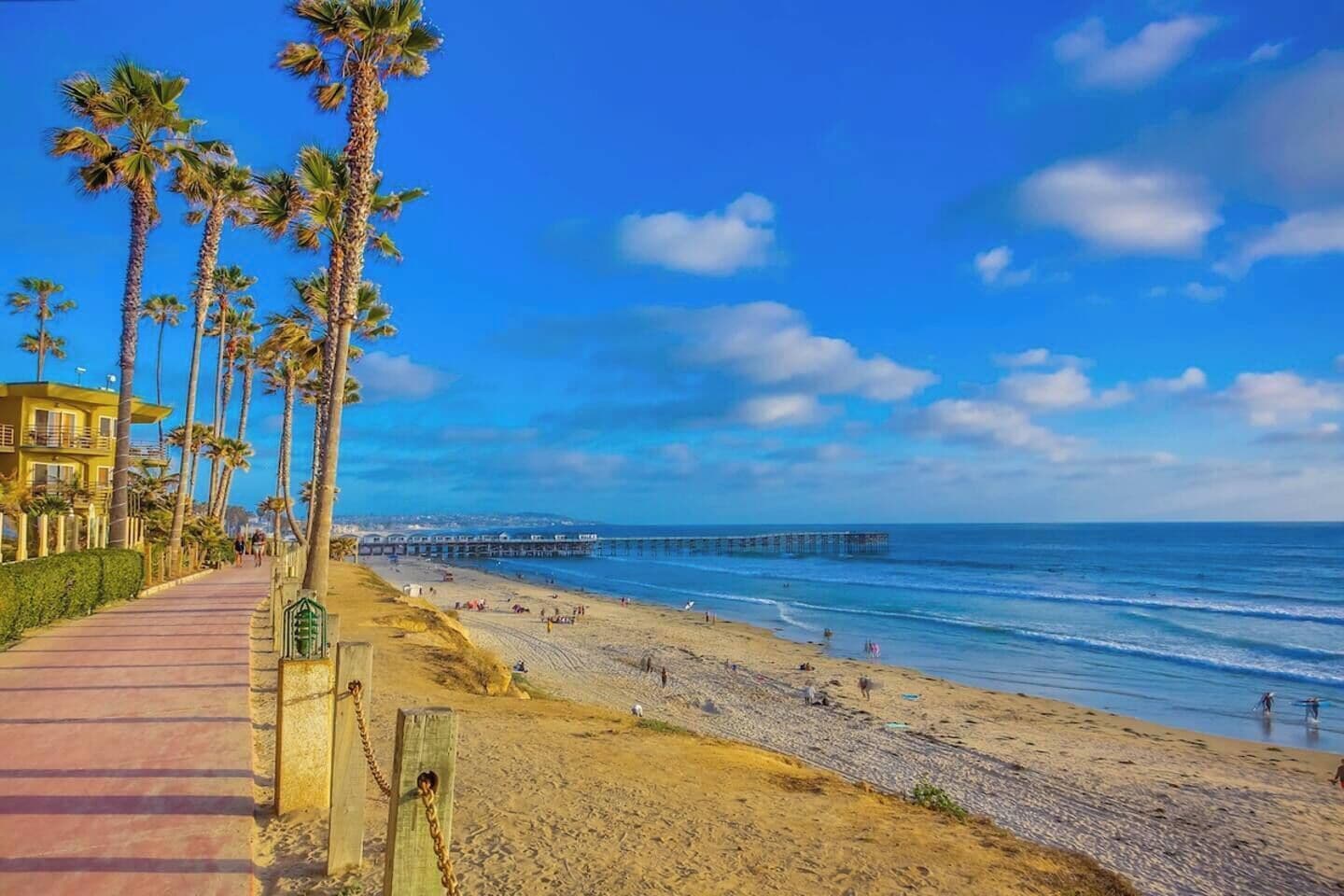 Ein Strand mit einer Holzpromenade, gesäumt von Palmen, einer Pier, die ins Meer hinausführt, und einem klaren, blauen Himmel.