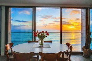 Un'area pranzo con un tavolo rotondo e sedie, un vaso con fiori rossi e un'ampia finestra vista oceano e tramonto.