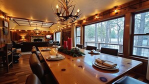 A dining area with a long wooden table set for four, a fireplace, and large windows offering a view of trees.