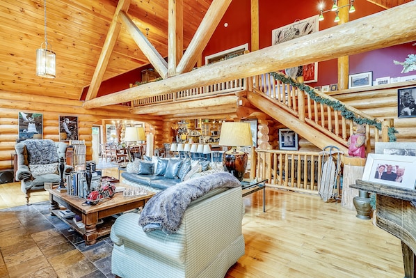 Interior de una cabaña de troncos con una escalera de madera, una sala de estar con sofá y sillones, y un comedor.