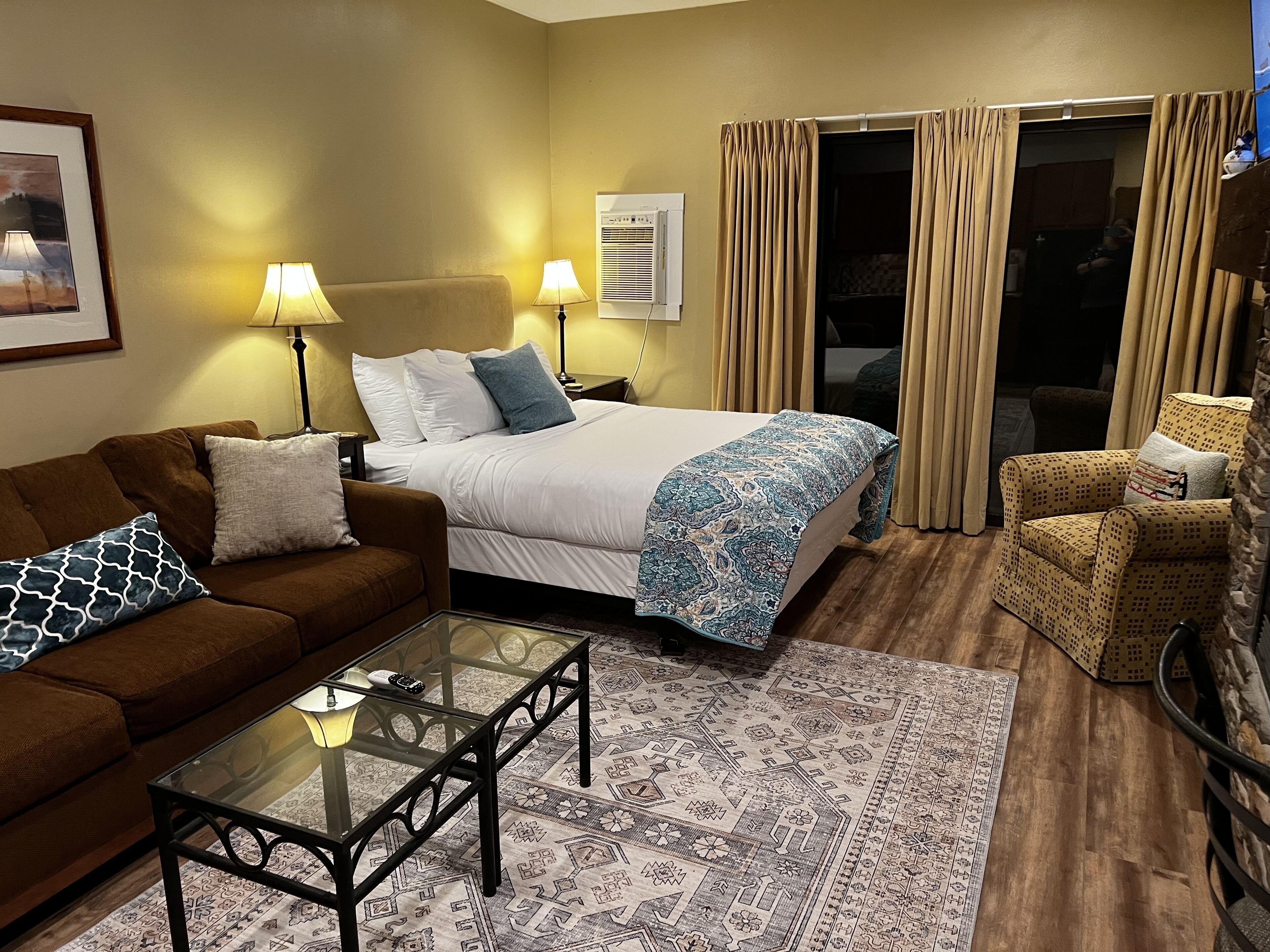 Quarto de hotel com cama, sofá, mesa de centro de vidro e tapete estampado.