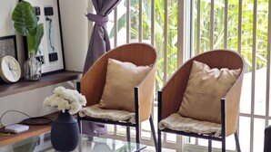 A modern living room with two wooden chairs, a glass coffee table, and a view of lush greenery through large windows.