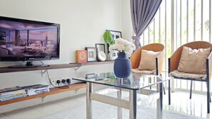 A modern living room with a flat-screen TV, a glass coffee table, a bookshelf, and two chairs with cushions.
