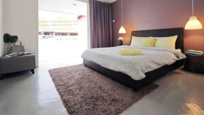 A modern bedroom with a large bed, a grey armchair, a small table with a vase, and a view of the outside.