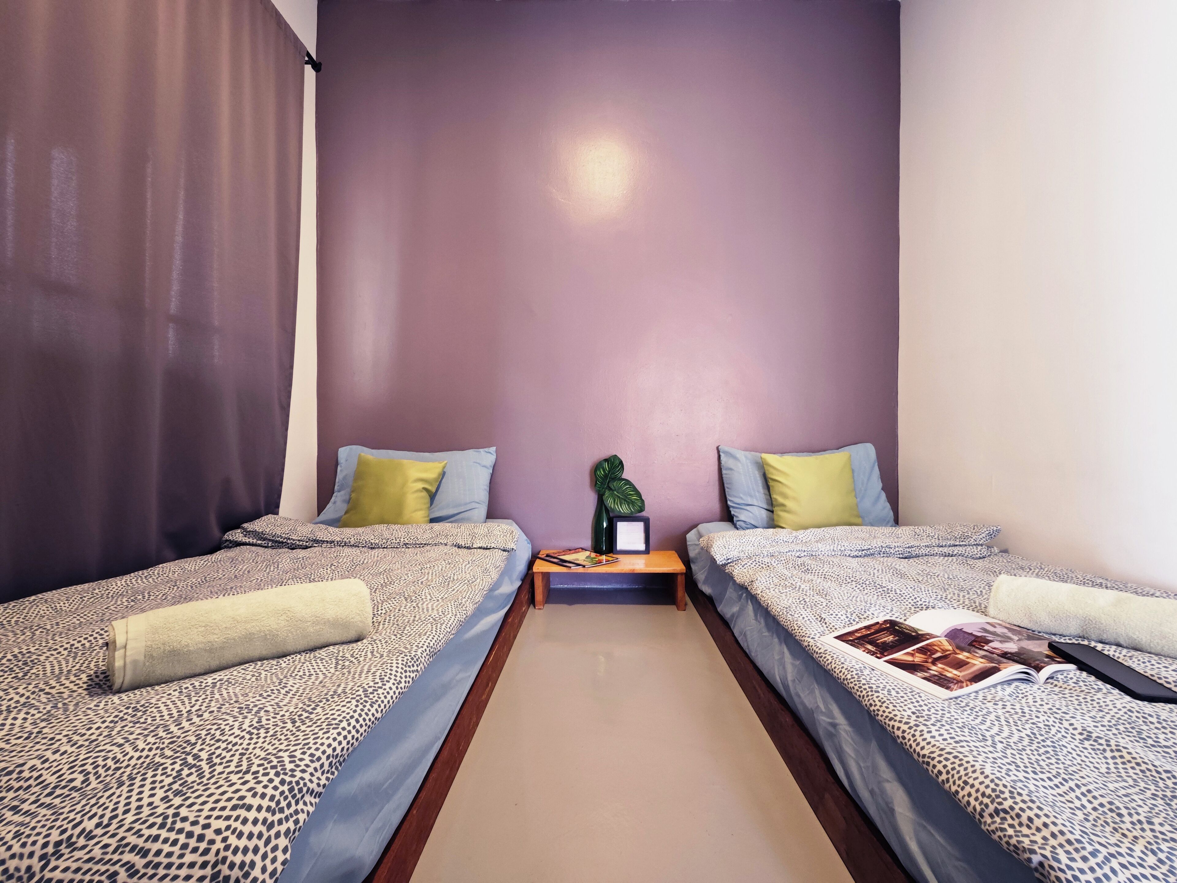 A small, well-lit room with two single beds, each with a patterned bedspread and pillows. There is a nightstand with a lamp and a small plant between the beds.
