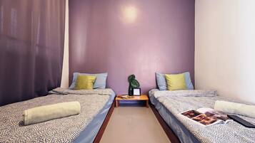 A small, well-lit room with two single beds, each with a patterned bedspread and pillows. There is a nightstand with a lamp and a small plant between the beds.
