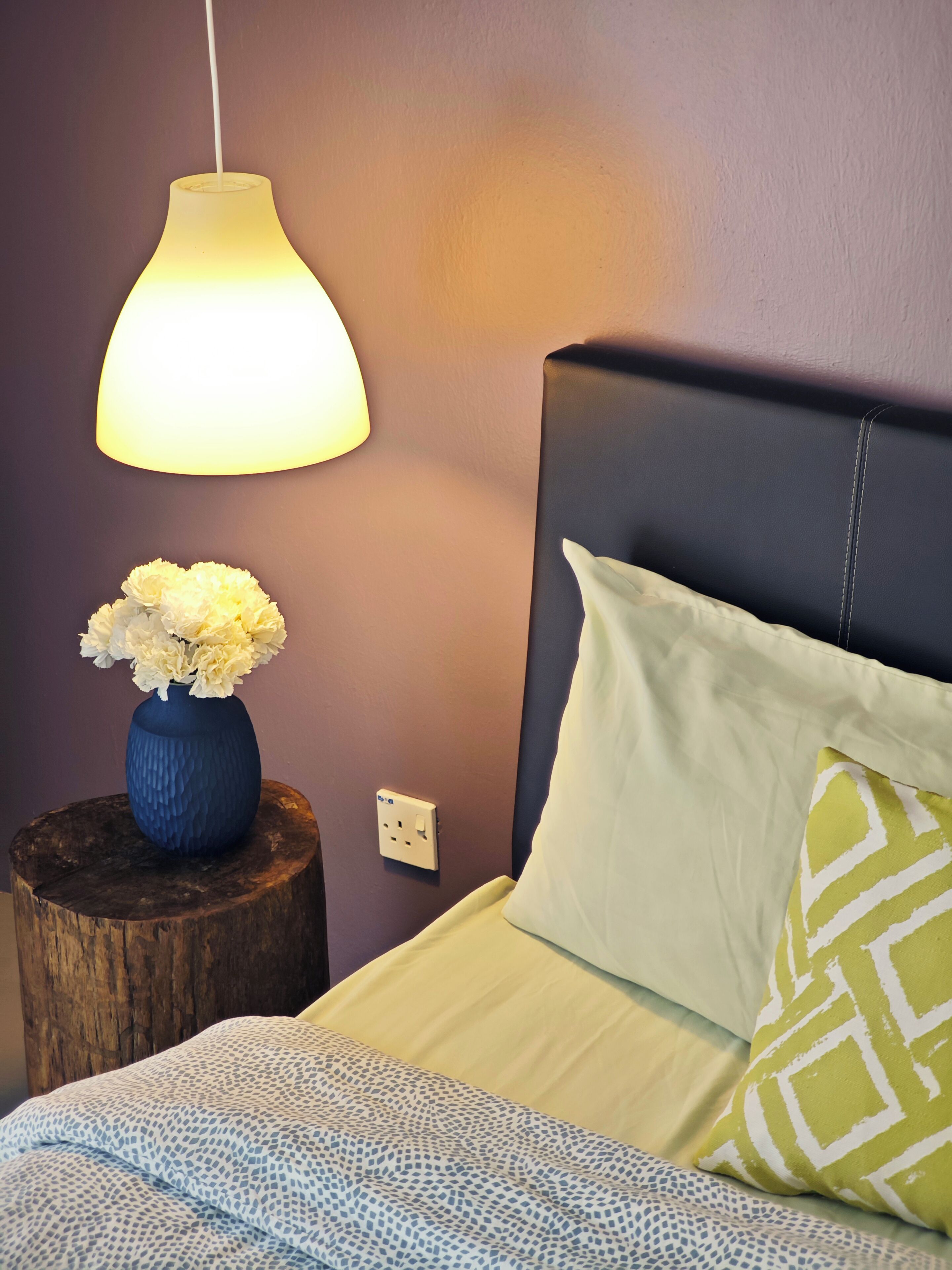 A bedroom with a bed, a bedside table with a vase of flowers, a lamp, and a power outlet on the wall.