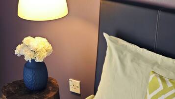 A bedroom with a bed, a bedside table with a vase of flowers, a lamp, and a power outlet on the wall.