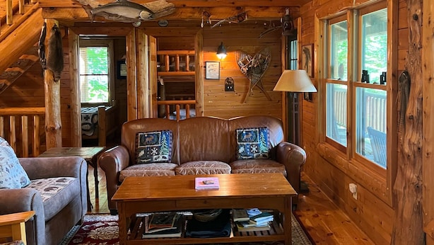 A cozy cabin interior with a wooden staircase, a leather sofa with patterned cushions, a red floral carpet, and a wooden table. - A beautiful log cabin home on Lake Huron, amazing lake views, hot tub, game room (Cheboygan)