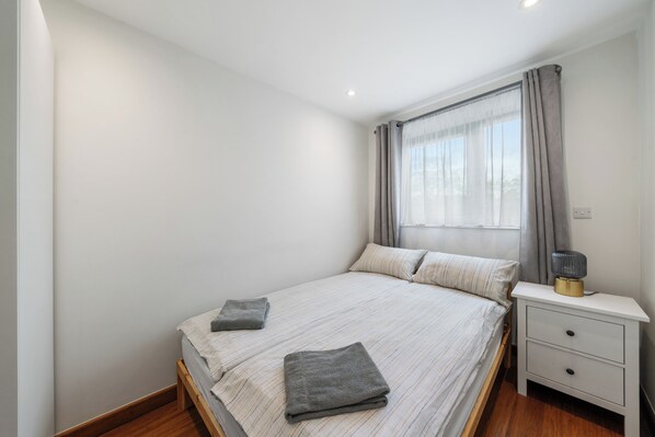 A bedroom with a bed, two pillows, a bedside table with a lamp, and a window with curtains.