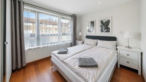 Un dormitorio moderno con dos camas, un ventanal con cortinas, una mesita de noche con lámpara y una obra de arte en la pared.