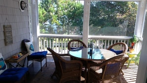 A sunlit patio with wicker furniture, a glass table, and a view of trees through a screened window.