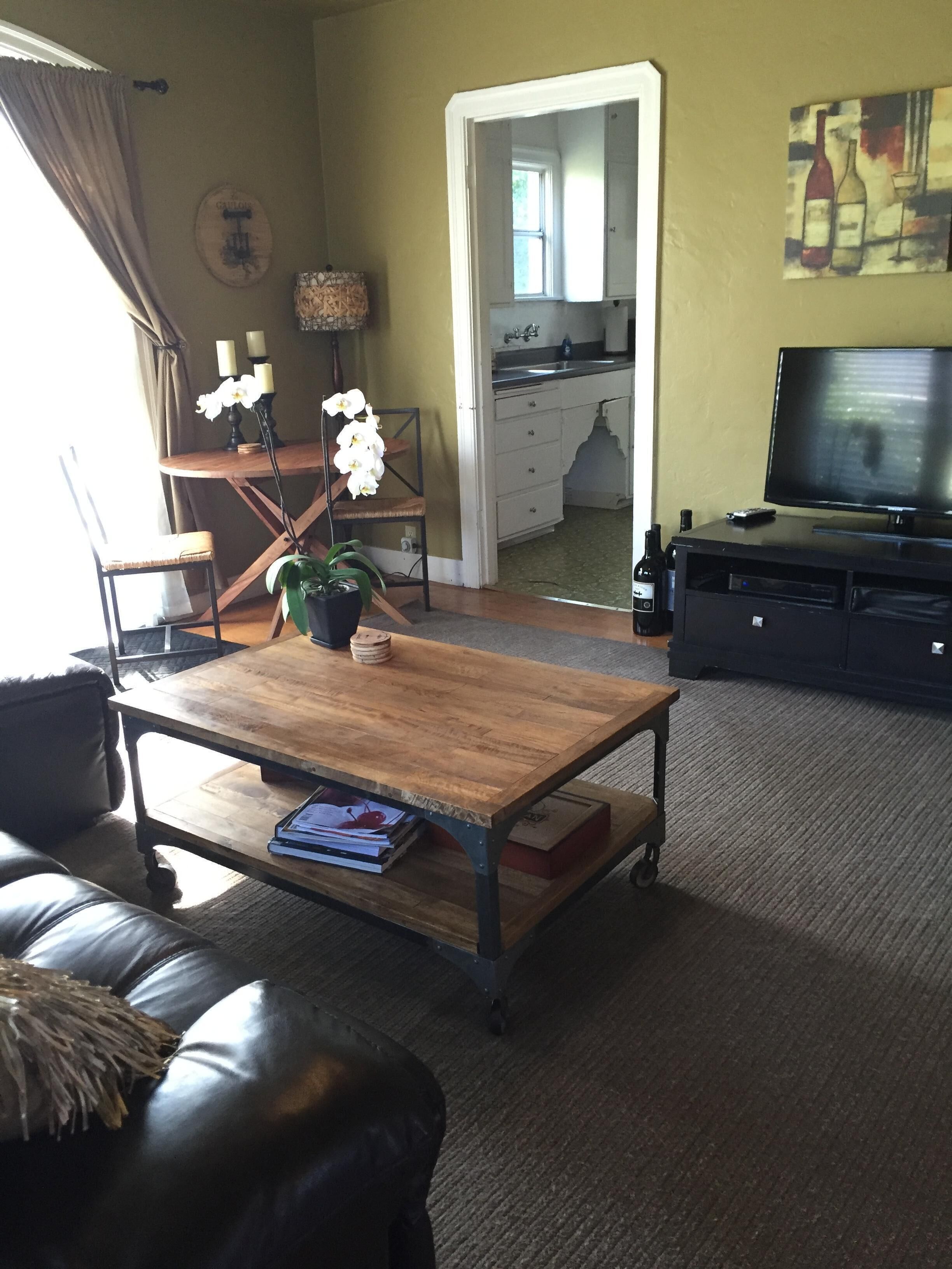 Ein Wohnzimmer mit Ledersofa, Couchtisch aus Holz, Fernseher und einer durch einen offenen Durchgang sichtbaren Küche.