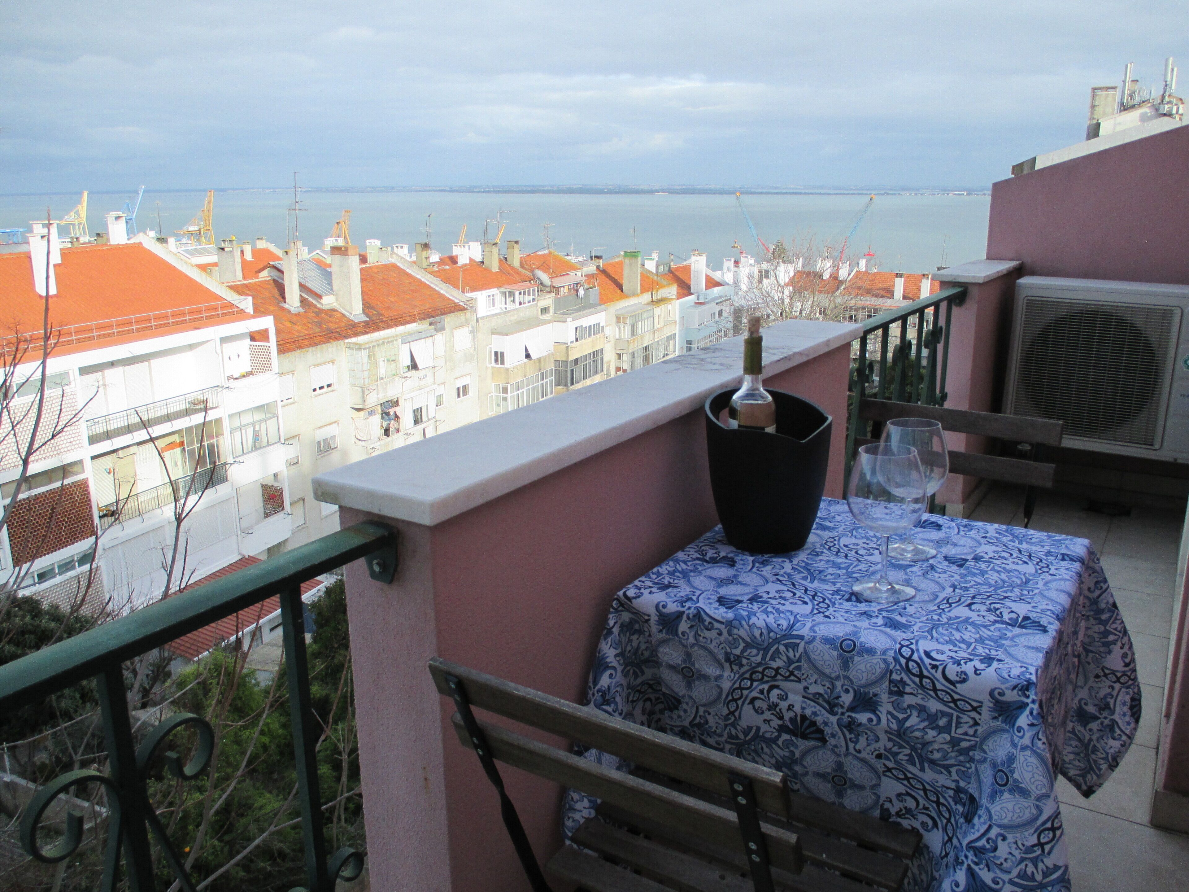 A balcony with a table set for two, overlooking a cityscape with red-roofed buildings and a body of water.