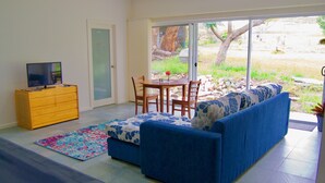 A living room with a sofa, a TV on a wooden cabinet, a dining table with chairs, and a sliding glass door leading to a garden. - Back Valley Farmstay Bed and Breakfast (Back Valley)