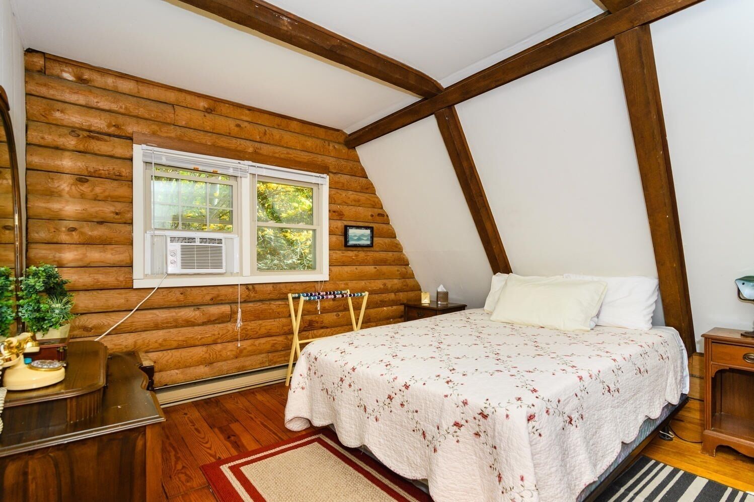A bedroom with a sloped ceiling, wooden walls, a bed with a floral bedspread, a small bedside table, and a window with an air conditioning unit.