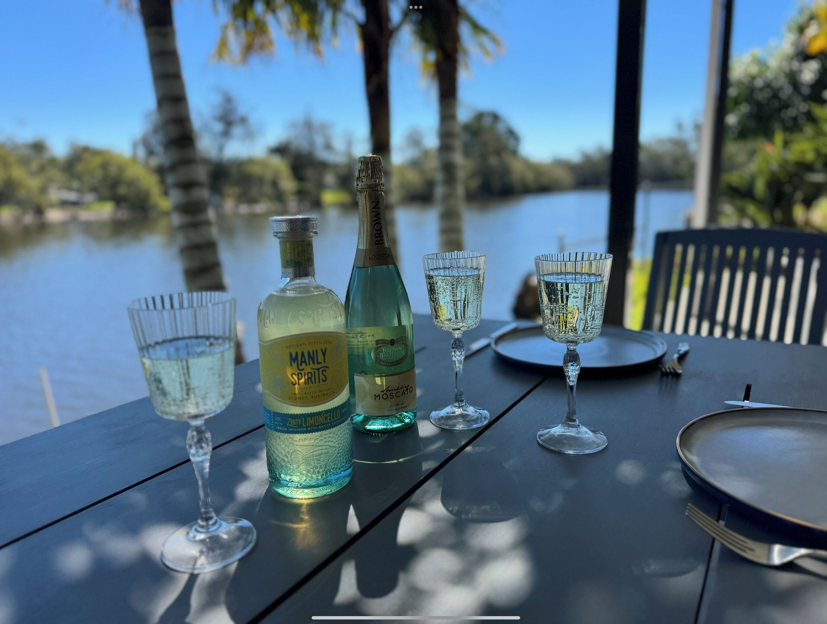 A table with a bottle of Manly Spirits, a bottle of Moscato, and three glasses of water, set against a lakeside view.
