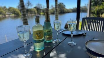 A table with a bottle of Manly Spirits, a bottle of Moscato, and three glasses of water, set against a lakeside view.