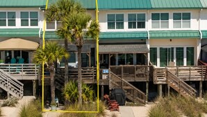 Une propriété située en bord de mer, avec un bâtiment surmonté d’un toit végétalisé, un espace extérieur couvert aménagé en coin salon et un escalier en bois menant à une terrasse.