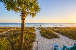 A beach pathway leading to a wooden deck with chairs, surrounded by sand and grass.