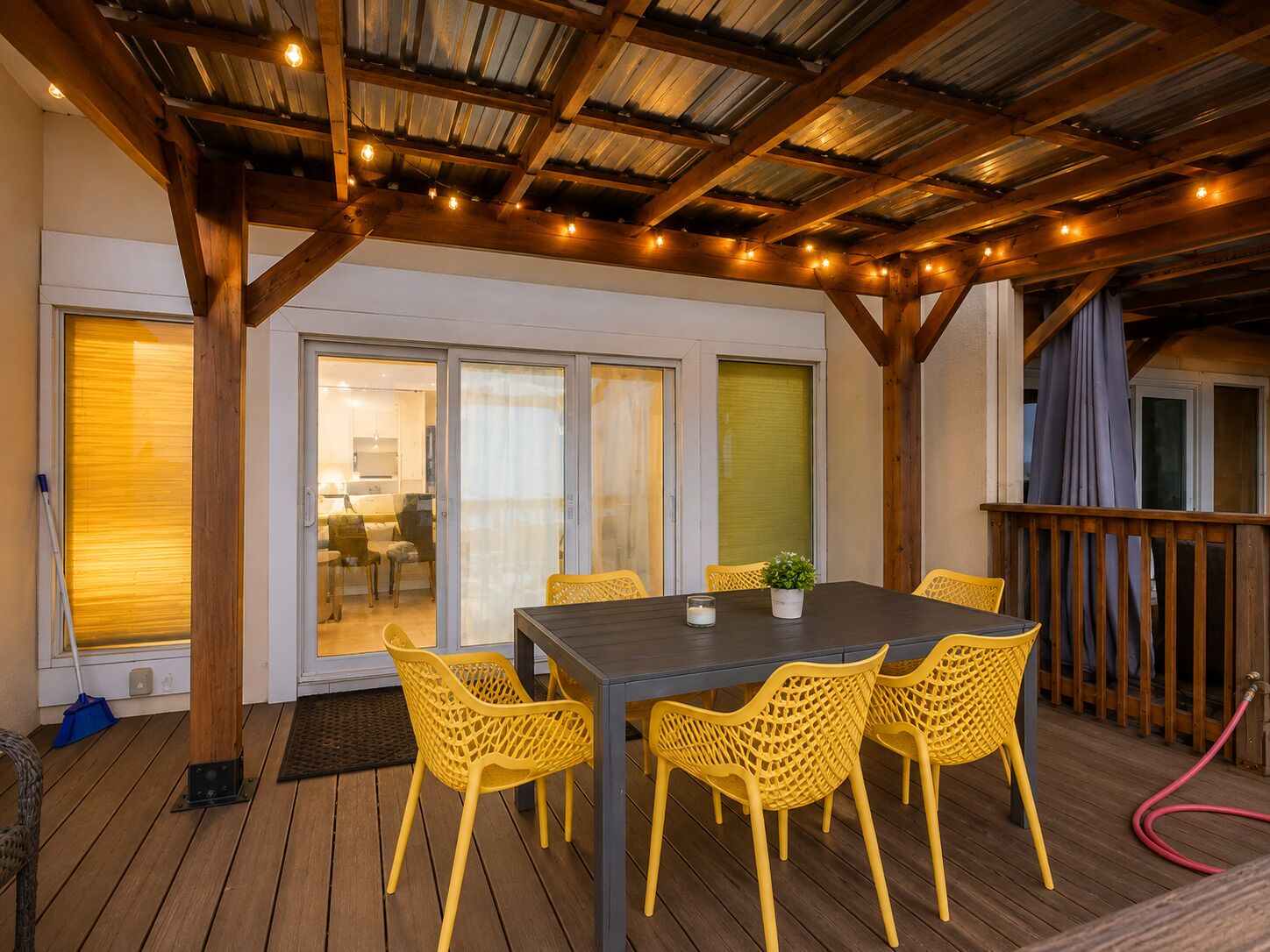 Um pátio externo com deck de madeira, cadeiras amarelas, uma mesa e área coberta com corrimão.