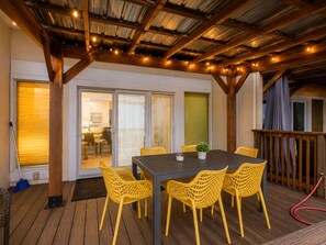 An outdoor patio with a wooden deck, yellow chairs, a table, and a covered area with a railing.