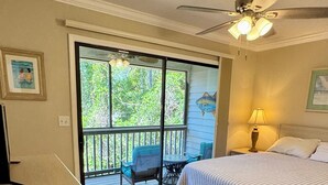Un dormitorio con cama, escritorio, silla, balcón y ventilador de techo, y vistas a árboles.