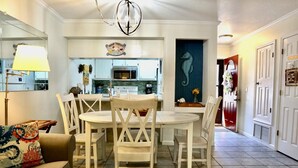 Un comedor con una mesa redonda y sillas blancas, una cocina con armarios azul claro, y un pasillo con un cuadro de un caballito de mar.