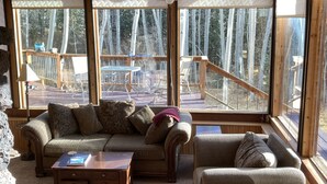 A cozy cabin interior with a wooden ceiling, large windows, a sofa, a coffee table, and a view of a wooden deck and trees outside.