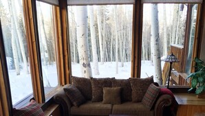 A cozy living room with a sofa, armchairs, and a coffee table. Large windows offer a view of a snowy forest.