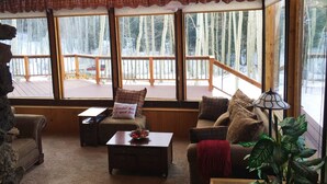 A cozy cabin interior with a wooden deck, large windows with a view of trees, and a fireplace.
