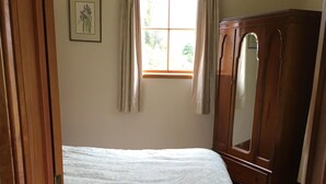 Un dormitorio con suelo de madera, una cama con cubrecama blanco, un armario de madera, una ventana con cortinas y un cuadro enmarcado en la pared.