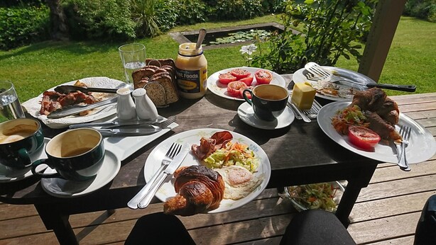 Una mesa de madera al aire libre con diversos alimentos para el desayuno, como huevos, tocino y un croissant.