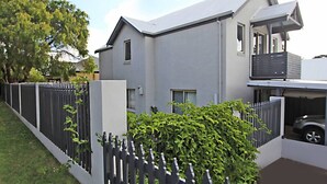 Une maison moderne à deux étages, avec une façade grise, un balcon à l’étage supérieur et une cour clôturée agrémentée de verdure.