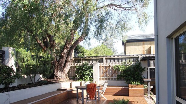 Une terrasse en bois avec une table et des chaises, entourée d’un mur blanc et d’un grand arbre.