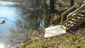 Una escalera de madera que desciende hacia la orilla de un río, rodeada de árboles y una zona cubierta de césped.