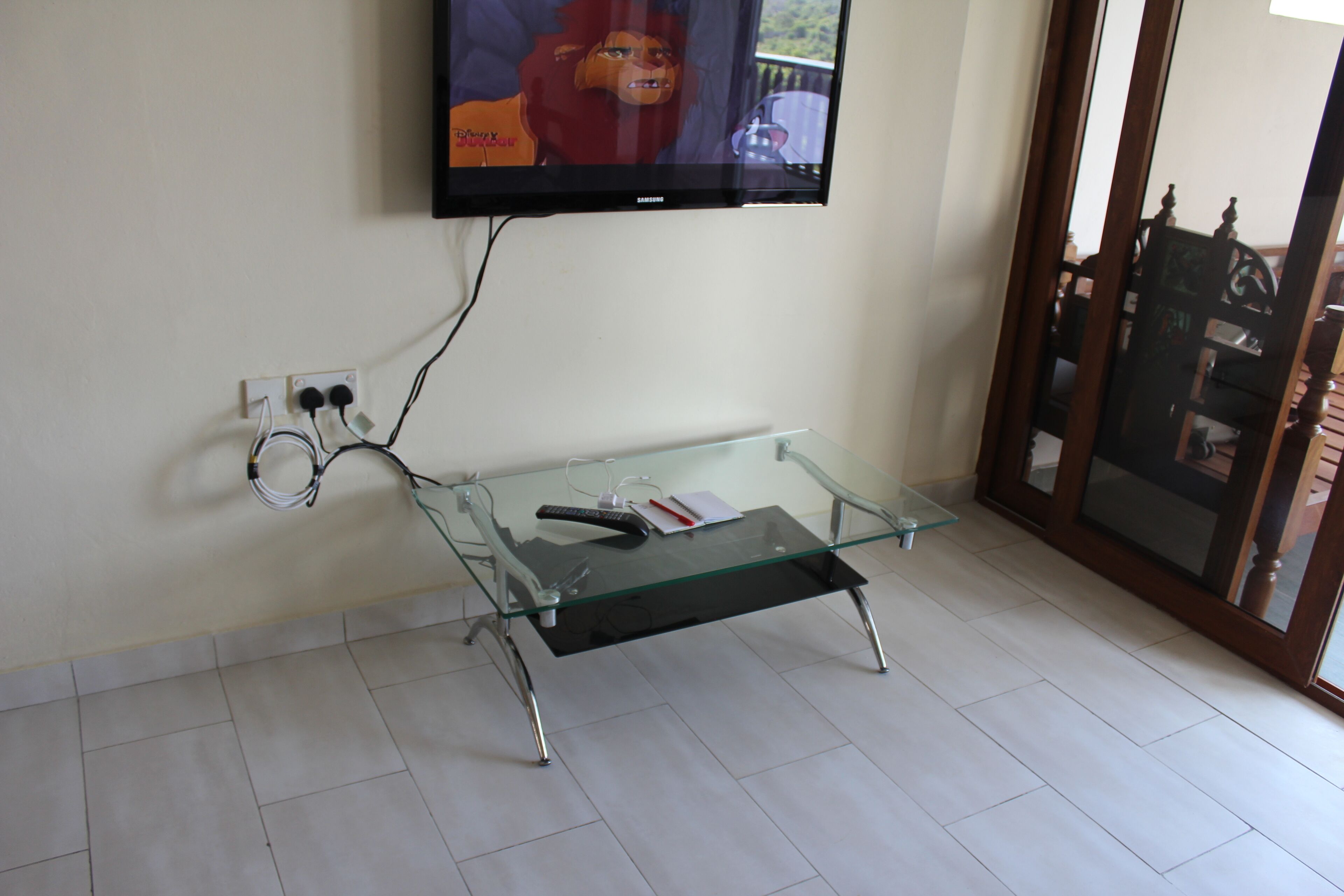 A glass-top coffee table with a remote control and a book, a wall-mounted TV, and a power outlet with cables.