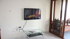 A living room with a flat-screen TV mounted on the wall, a glass coffee table with a remote control, and a wooden door with glass panels. - Beach Front Apartment. (Diani Beach)
