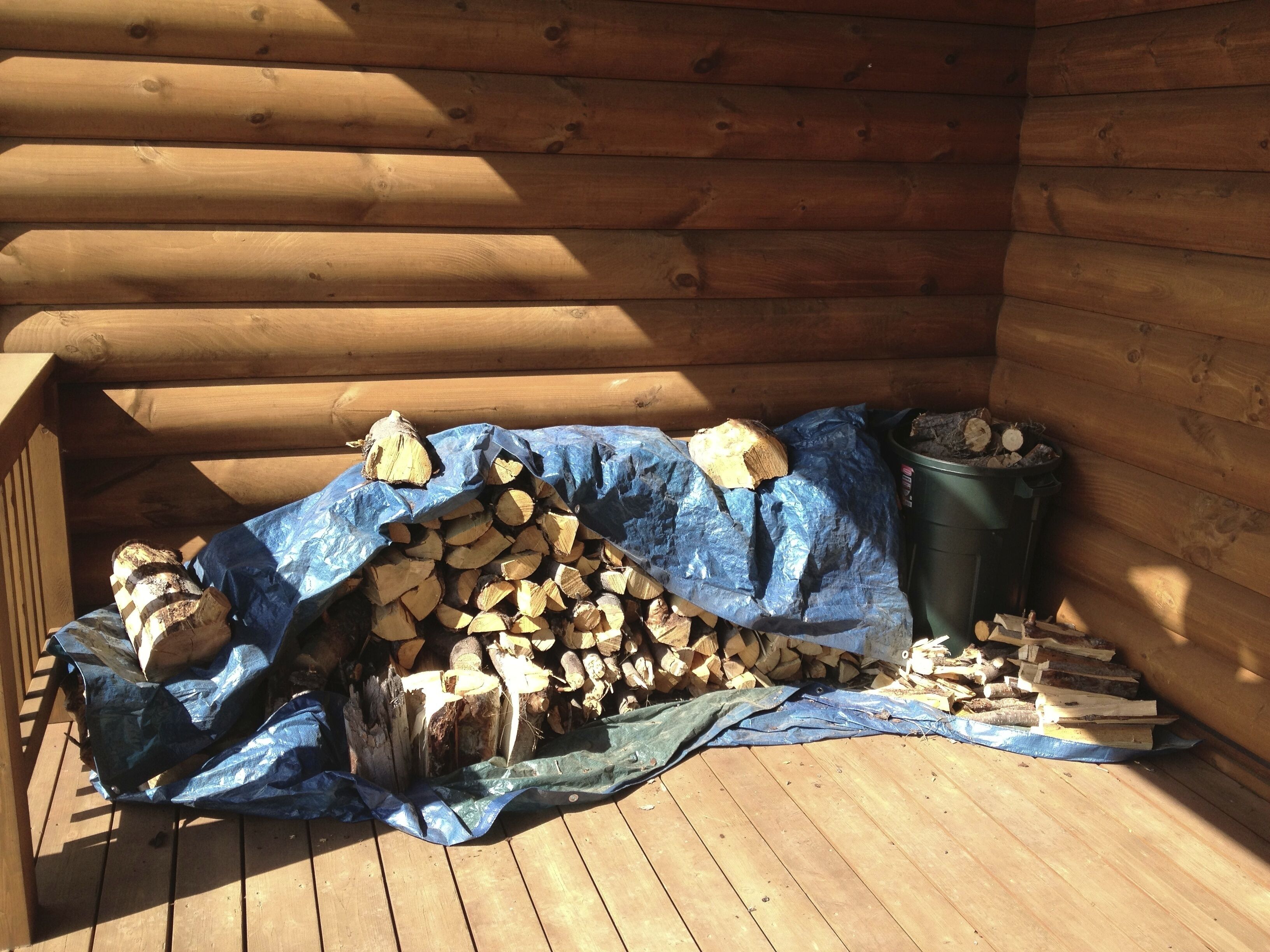 A wooden cabin with a pile of firewood and a green trash bin on the deck.