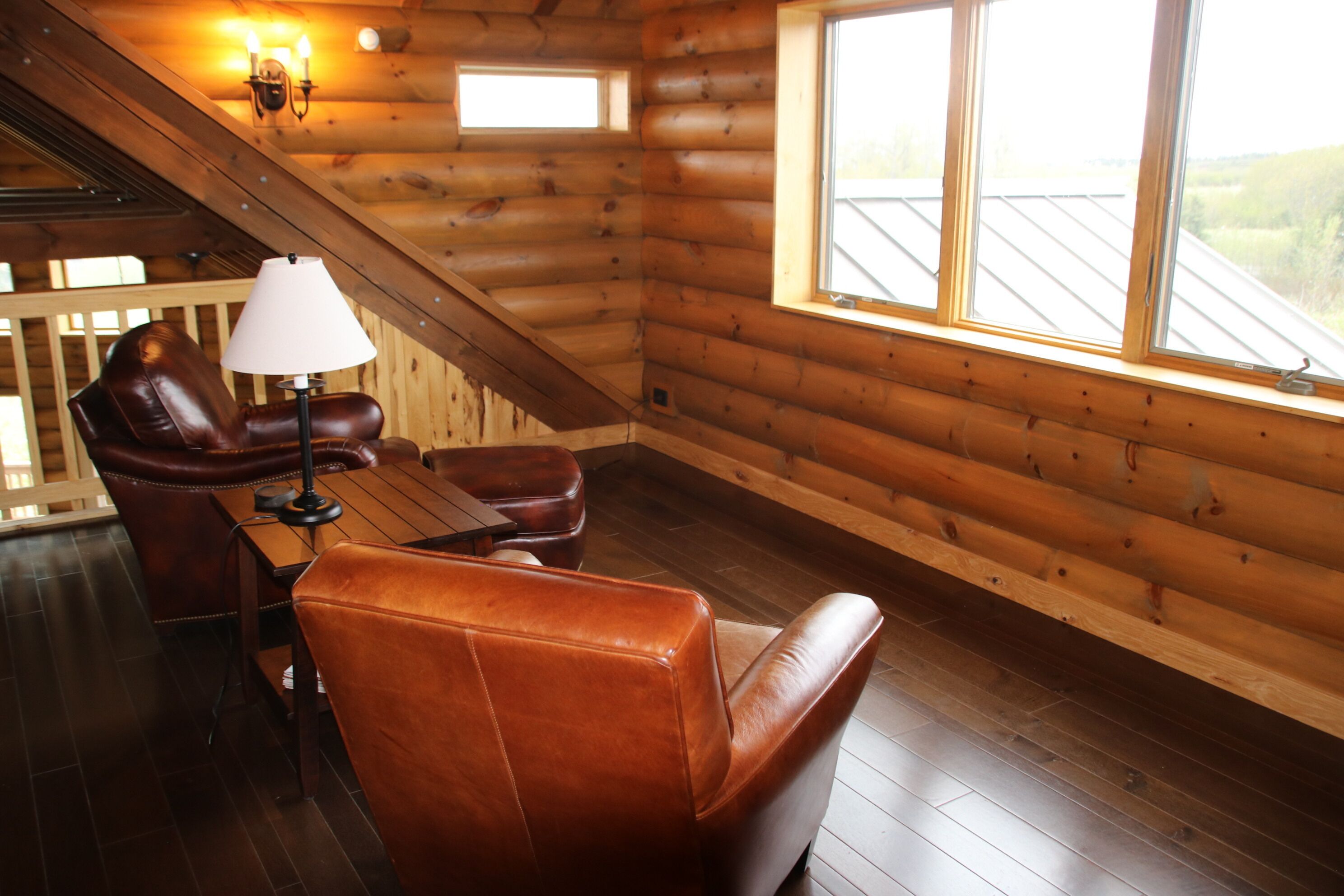 A cozy log cabin room with a leather sofa, a small table, and a lamp. There are two windows providing natural light and a view of the outdoors.