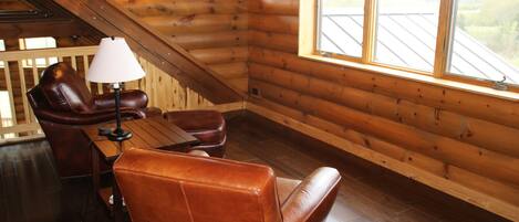 A cozy log cabin room with a leather sofa, a small table, and a lamp. There are two windows providing natural light and a view of the outdoors.