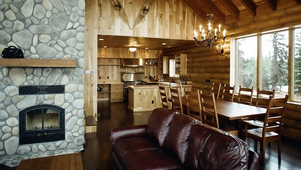 A log cabin interior with a stone fireplace, wooden dining table, and a leather couch.