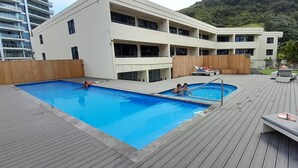 A swimming pool with a wooden deck, a building with balconies, and a mountainous backdrop.