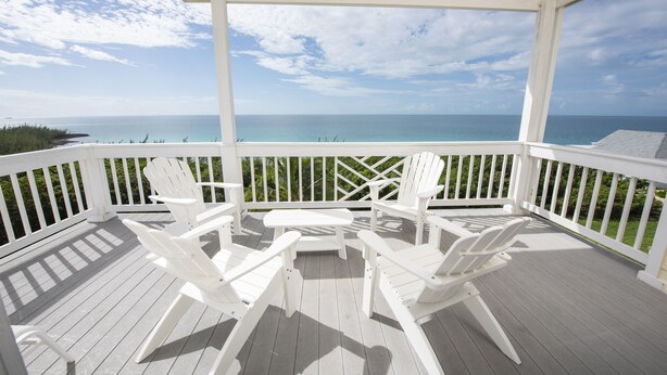 A covered deck with white Adirondack chairs and a table, overlooking a clear blue ocean. - Condo w/Views, Beach, Htd Pool, Pickleball, Golf Cart, Gym,Walk to Restaurant (Governors Harbour)