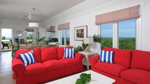 A spacious living area with red sofas, a white coffee table, and large windows offering a view of the ocean. - Condo w/Views, Beach, Htd Pool, Pickleball, Golf Cart, Gym,Walk to Restaurant (Governors Harbour)