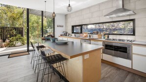 A modern kitchen with a central island, bar stools, a large window, and a view of greenery. - Tucker House (Wye River)