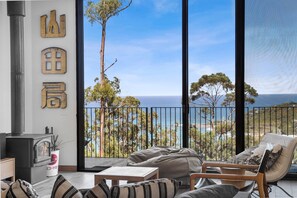 A modern living room with a large window overlooking a coastal view, a fireplace, and a balcony with a railing. - Tucker House (Wye River)