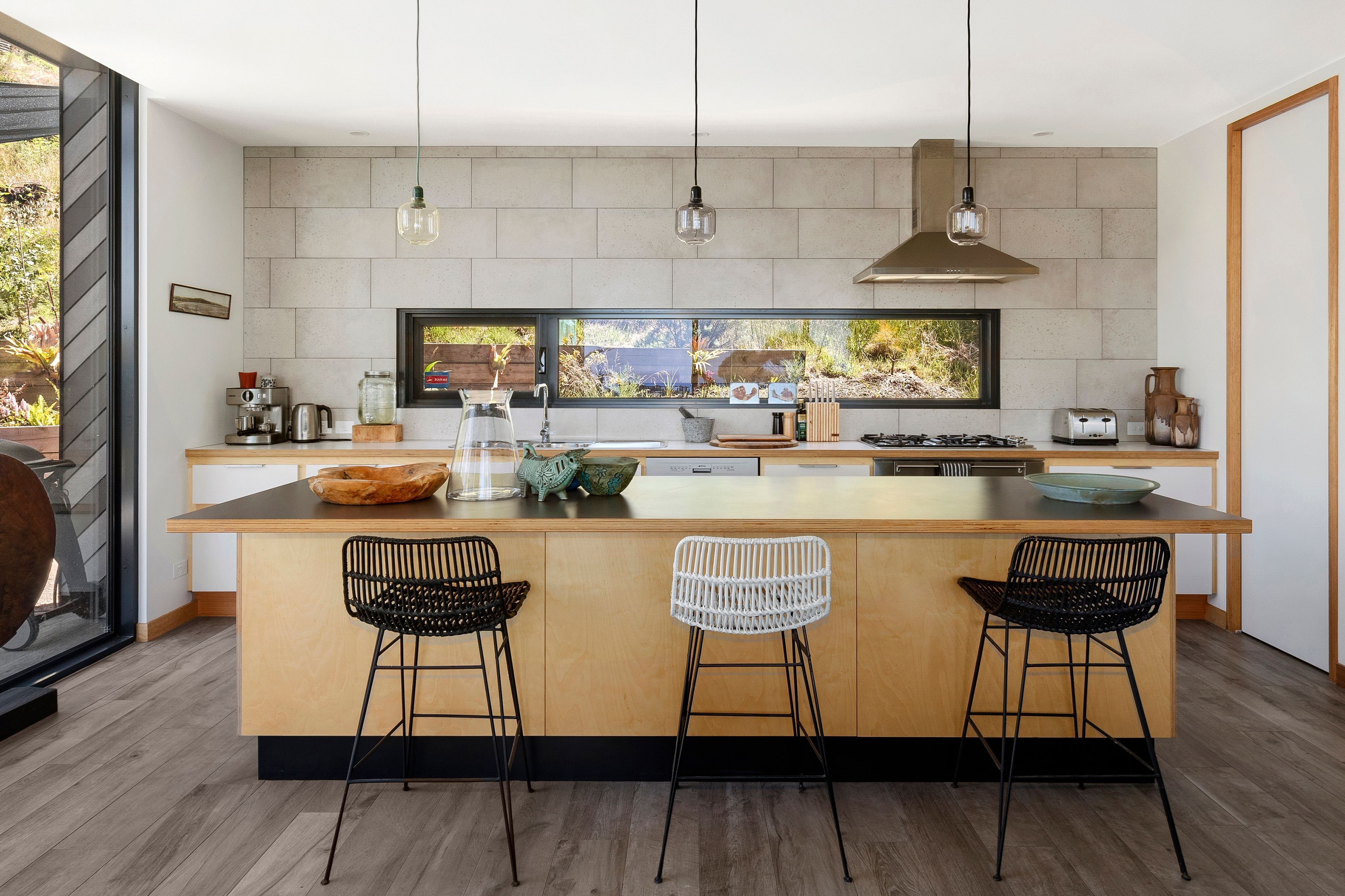 A modern kitchen with a large island, bar stools, a coffee machine, and a view through the window.